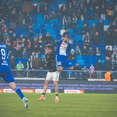 Na zdjęciu widoczna jest akcja podczas meczu piłkarskiego, gdzie zawodnik skacze w powietrze, a w tle widać zebranych kibiców na trybunach.