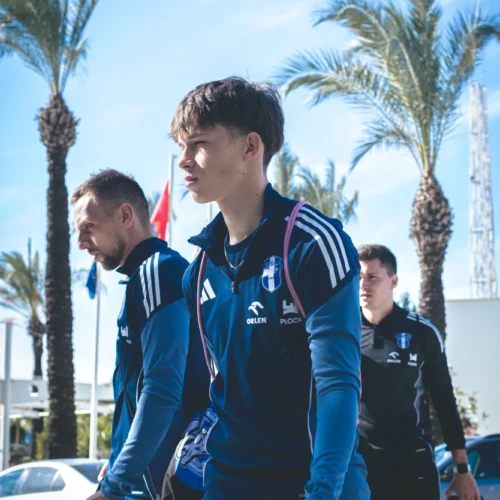 Trzech młodych sportowców w odzieży treningowej niesie buty, w tle widać palmy i błękitne niebo.