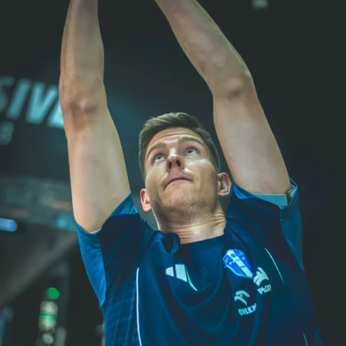 Mężczyzna w niebieskim t-shircie wykonuje ćwiczenie siłowe, trzymając ciężar nad głową.