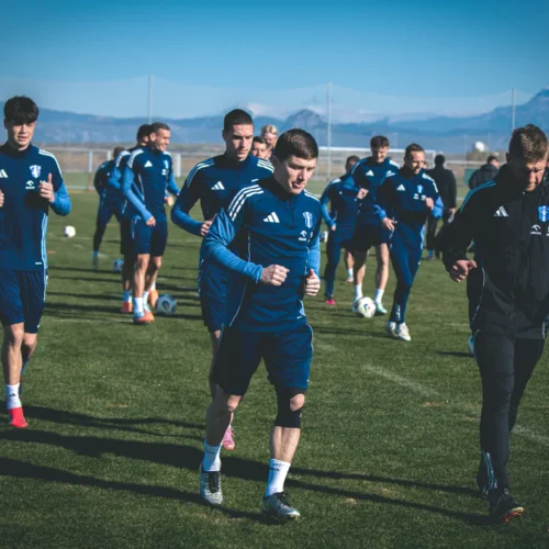 Piłkarze w niebieskich strojach trenują na boisku w słoneczny dzień, biegając w grupie.