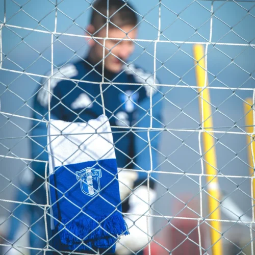 Na zdjęciu widoczny jest bramkarz, który stoi za siatką bramki, a na niej powiewa szalik w kolorach klubu Wisła Płock.