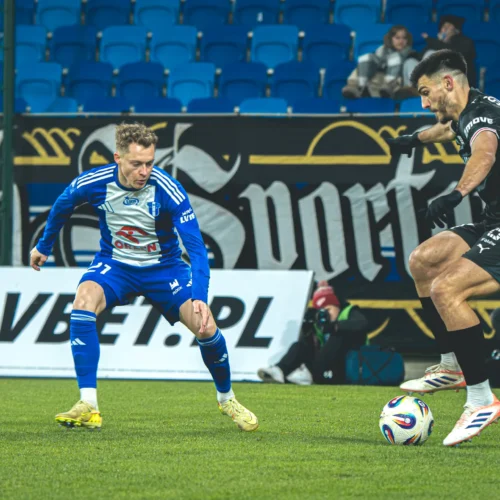 Na zdjęciu dwóch piłkarzy podczas meczu, zmierzających do piłki na murawie, w tle widoczne niebieskie krzesełka stadionowe oraz reklamy.