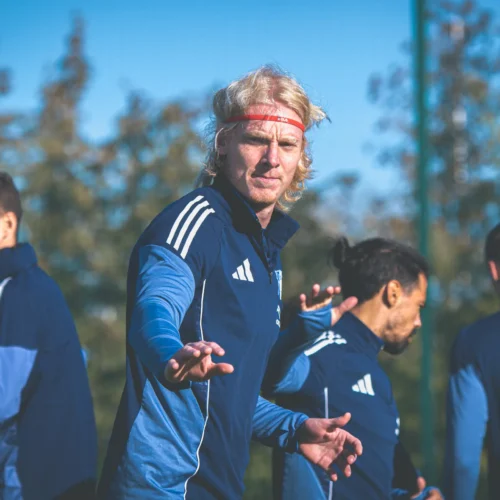 Na zdjęciu widać grupę piłkarzy trenujących na boisku w słoneczny dzień, z jednym zawodnikiem, który zwraca uwagę na widza.