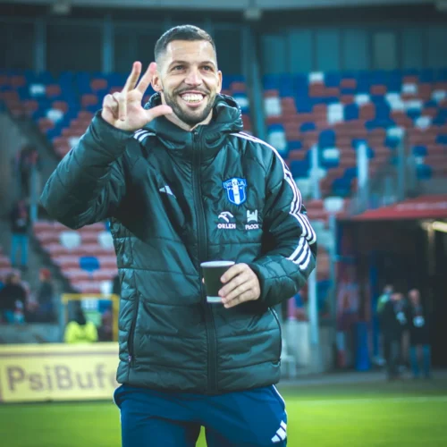 Na zdjęciu widać uśmiechniętego mężczyznę w sportowej kurtce, który trzyma kubek i wykonuje gest na znak pokoju.