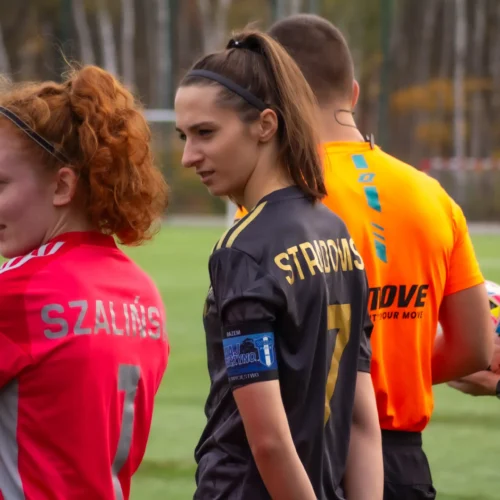 Na zdjęciu dwie zawodniczki drużyny piłkarskiej, jedna w czerwonym stroju z nazwiskiem "Szałyniś", a druga w czarnym z nazwiskiem "Stradows", stoją na boisku przed rozpoczęciem meczu.