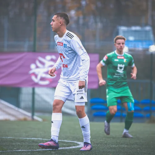 Na zdjęciu dwóch piłkarzy podczas meczu na boisku, jeden z nich w białym stroju, a drugi w zielonym.