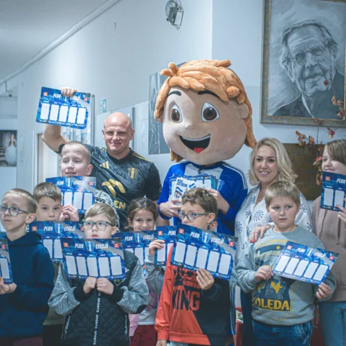 Grupa dzieci, dorosłych oraz maskotka z uśmiechem stoi w korytarzu, trzymając w rękach plansze do gry.