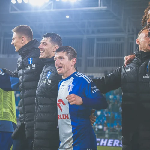 Na zdjęciu widoczna jest grupa radosnych piłkarzy świętujących zwycięstwo po meczu, uśmiechających się i trzymających się za ramiona w sportowych strojach.