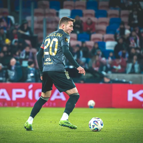 Na zdjęciu widoczny jest piłkarz w wyposażeniu sportowym, który przygotowuje się do zagrania piłki na boisku, z publicznością w tle.