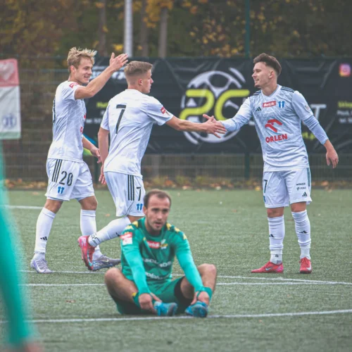 Na zdjęciu trzech piłkarzy w białych strojach radośnie świętuje na boisku, podczas gdy jeden z bramkarzy siedzi na murawie.