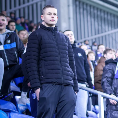 Grupa kibiców na stadionie, z wyraźnym fokusem na młodego mężczyznę w czarnej kurtce, stojącego na trybunach.