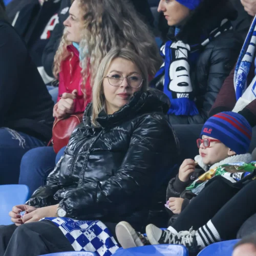 Na zdjęciu widać kobietę w czarnej kurtce siedzącą na trybunach z dzieckiem w niebieskiej czapce, które zajada się przekąskami.