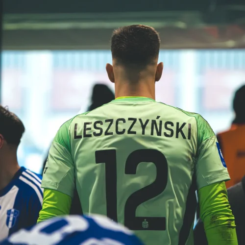 Na zdjęciu widać bramkarza w zielonej koszulce z numerem 12 oraz nazwiskiem "LESZCZYŃSKI", stojącego w szatni przed meczem.