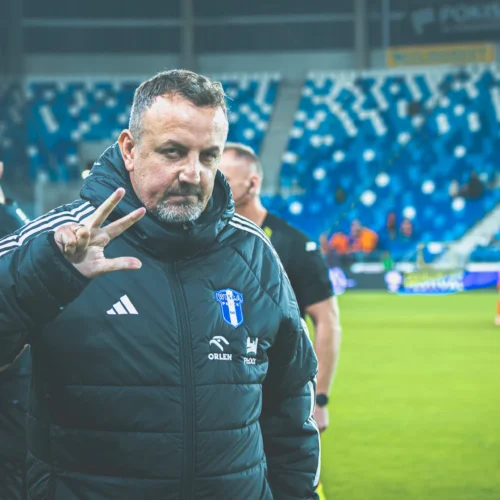 Na zdjęciu widoczny jest mężczyzna w czarnej kurtce, który wita się z kibicami, stojąc na boisku piłkarskim.