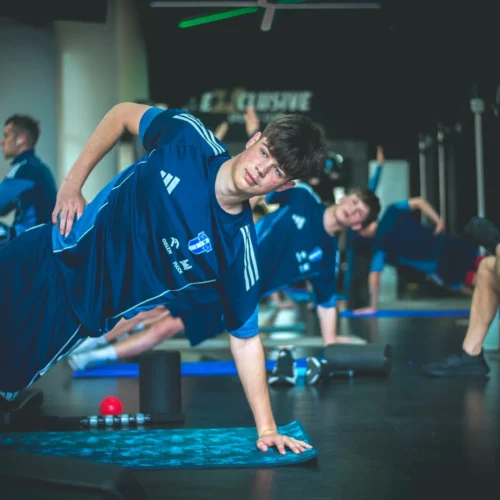 Na zdjęciu widać grupę młodych sportowców wykonujących ćwiczenia w siłowni, skupionych na poprawie swojej kondycji fizycznej.