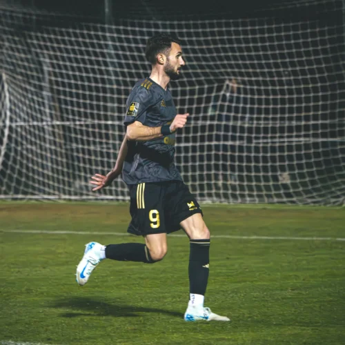 Na zdjęciu widać piłkarza w czarnej koszulce z numerem 9, który biegnie w kierunku bramki, a w tle znajduje się siatka bramkowa oraz bramkarz w żółtym stroju.