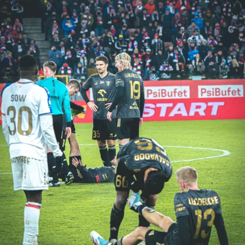 Na zdjęciu widoczna jest sytuacja na boisku piłkarskim, gdzie kilku zawodników otacza leżącego na ziemi kolegę z drużyny, a w tle widać kibiców na trybunach.