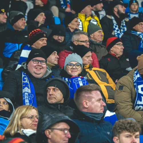 Na zdjęciu widać grupę kibiców na stadionie, z różnorodnymi wyrazami twarzy, ubranych w ciepłe odzież oraz szaliki w barwach drużyny.