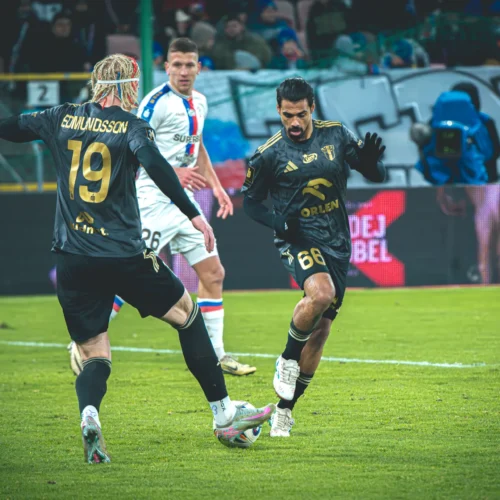 Na zdjęciu widać grupę zawodników piłkarskich podczas meczu, z jednym graczem w czarnej koszulce z numerem 66, który wykonuje drybling, a drugi zawodnik w czarnej koszulce z numerem 19 stoi obok niego.