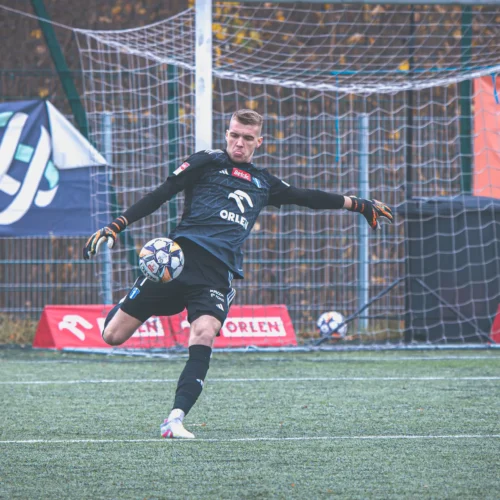 Bramkarz w czarnym stroju wykonuje interwencję na boisku piłkarskim, odbijając piłkę nogą.