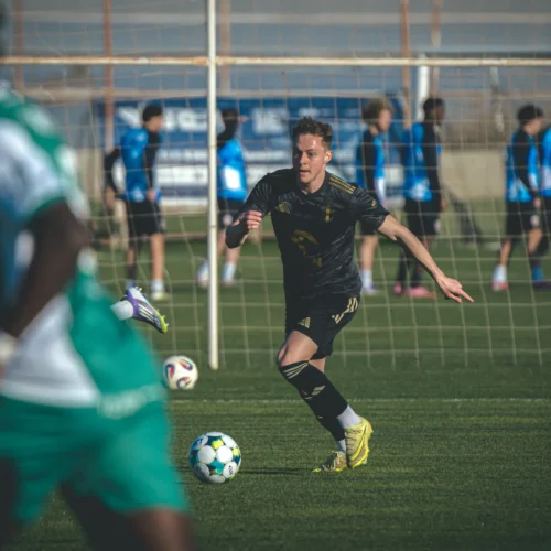 Młody piłkarz w czarnej koszulce biegnie za piłką na boisku, podczas gdy w tle trenują inni zawodnicy.
