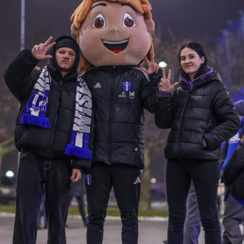 Trójka młodych ludzi, w tym jedna osoba w dużej pluszowej masce, pozują do zdjęcia na tle nocnego otoczenia, z wyciągniętymi palcami w geście pacyfikacji.