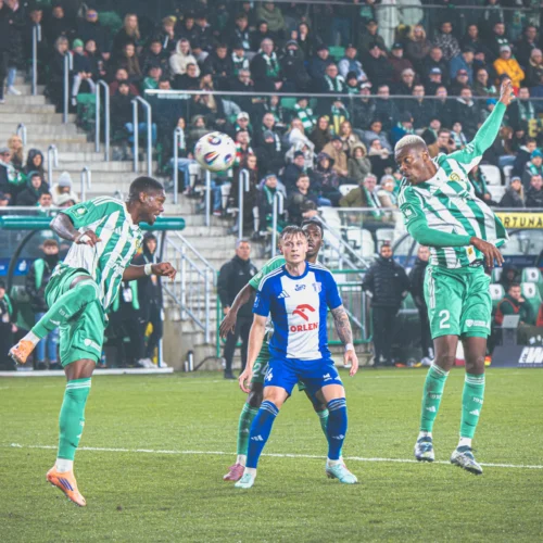 Na zdjęciu widoczni są zawodnicy drużyn piłkarskich w akcji na boisku, z jednym z graczy oddającym kontrolowane uderzenie piłki.