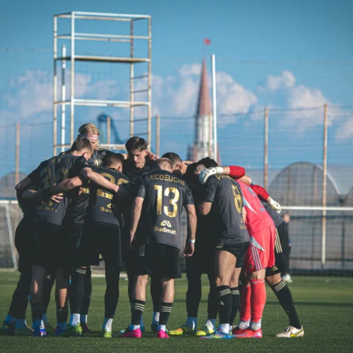Zdjęcie przedstawia zespół piłkarski w czarnych strojach, który zbiera się w okręgu przed meczem, na boisku w słoneczny dzień.