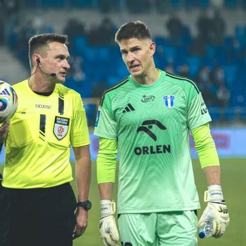 Na zdjęciu znajdują się sędzia w żółtym stroju oraz bramkarz w zielonym uniformie, rozmawiający na boisku podczas meczu piłkarskiego.