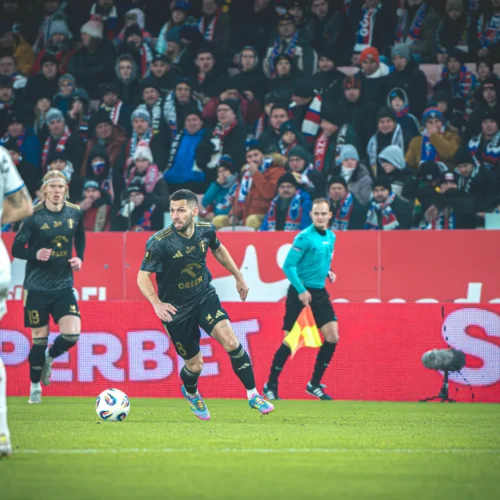 Na zdjęciu widać piłkarzy podczas meczu, w tle liczna grupa kibiców oraz sędzia na boisku.