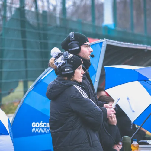 Na zdjęciu widoczni są dwoje ludzi w słuchawkach, obserwujących wydarzenie sportowe, z parasolami w rękach, w deszczowej aurze.