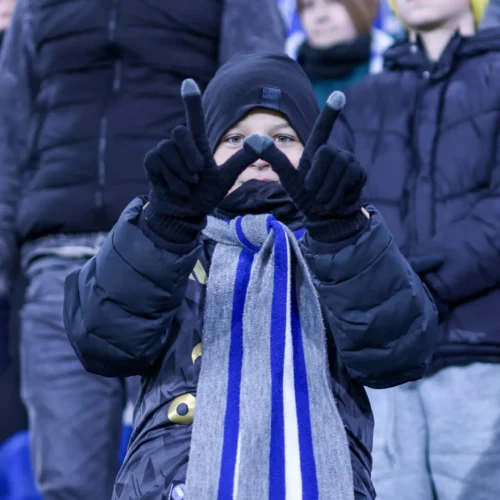Na zdjęciu widoczny jest mały chłopiec w zimowym ubraniu, który z entuzjazmem pokazuje znak V, trzymając w rękach szalik w kolorach szaro-niebieskich.