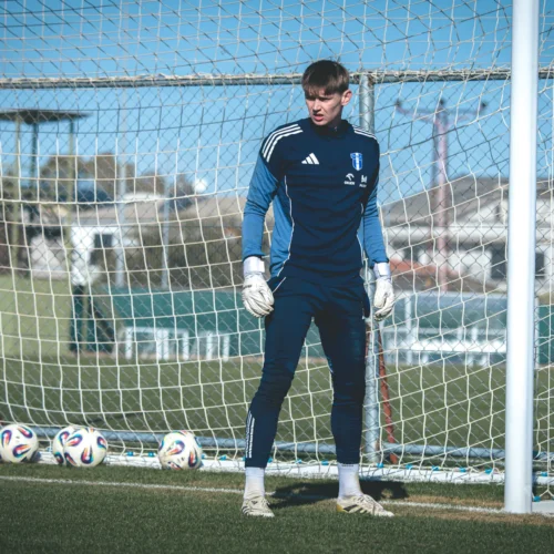 Na zdjęciu młody bramkarz w niebieskim stroju sportowym stoi w bramce na boisku piłkarskim, otoczony przez piłki.