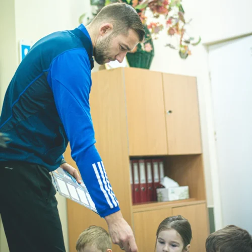 Mężczyzna w niebieskiej bluzie pokazuje coś dzieciom siedzącym przed nim w jasnym pomieszczeniu.