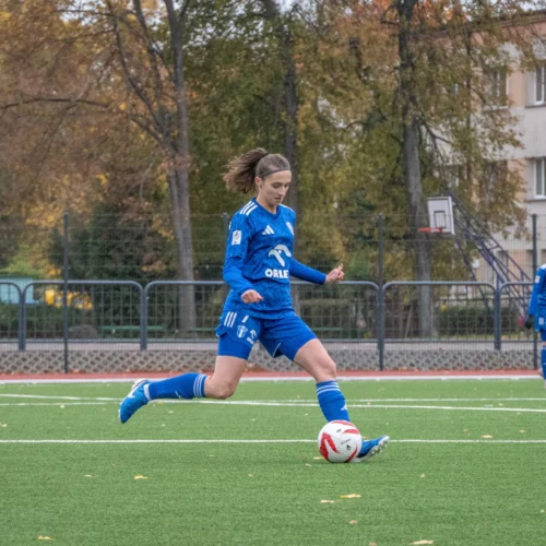 Na zdjęciu widoczna jest piłkarka w niebieskim stroju, która wykonuje strzał na boisku, podczas gdy jej koleżanki z drużyny obserwują akcję w tle.