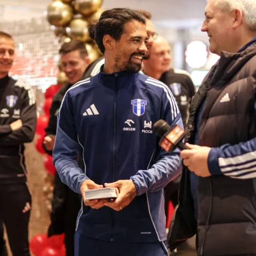 Na zdjęciu widać uśmiechniętego mężczyznę w niebieskim dresie, rozmawiającego z innym mężczyzną, podczas wydarzenia w towarzystwie kilku osób na tle ozdób świątecznych.