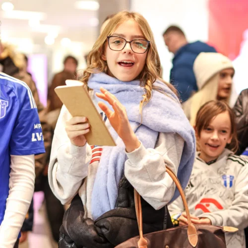 Na zdjęciu widać uśmiechniętą dziewczynkę trzymającą tablet, otoczoną przez innych ludzi w tle.