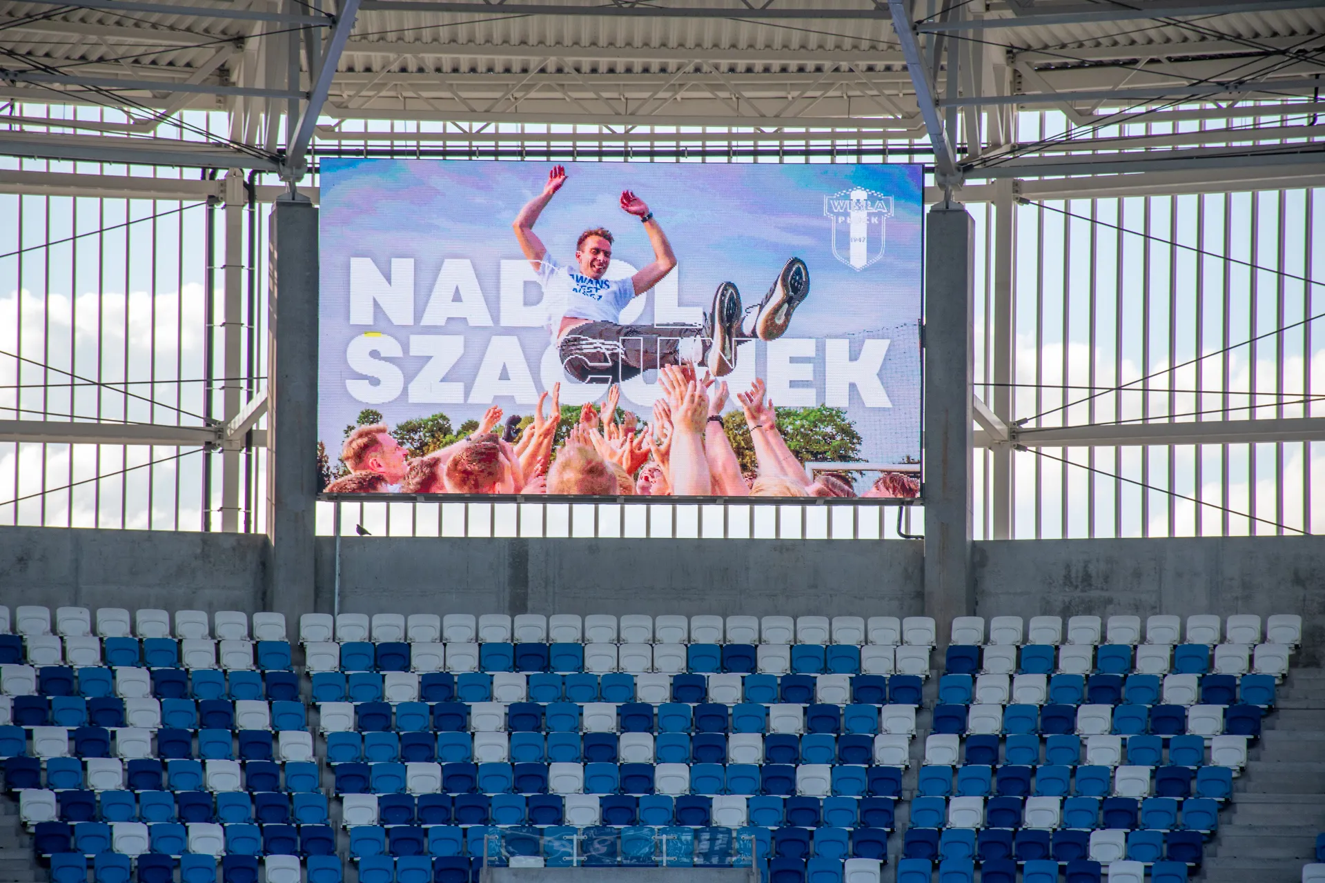 Na zdjęciu widać reklamę na wielkim ekranie, na której mężczyzna zostaje uniesiony przez tłum fanów, a obok napis