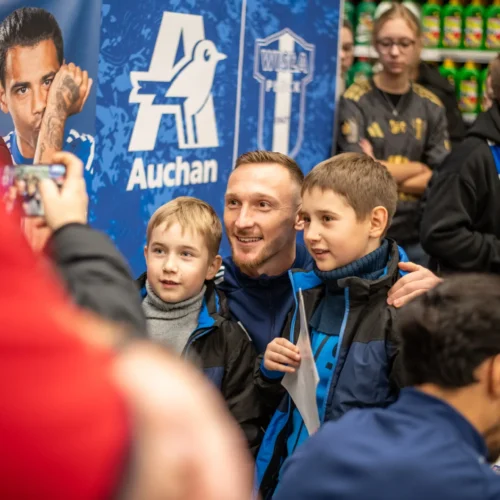 Na zdjęciu dwóch chłopców uśmiecha się do obiektywu razem z dorosłym mężczyzną, prawdopodobnie sportowcem, podczas wydarzenia w sklepie.
