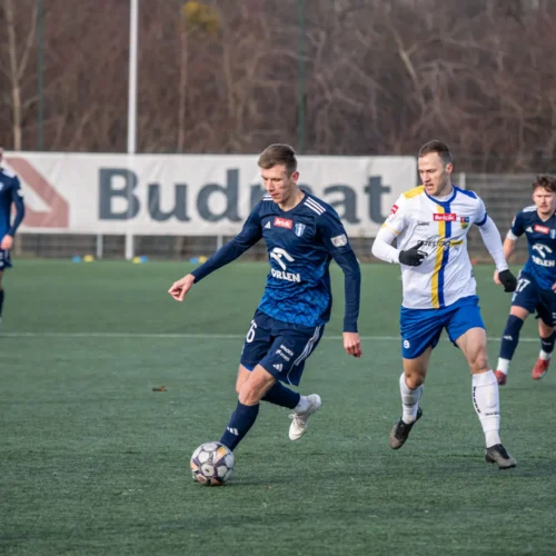 Na zdjęciu widać dwóch piłkarzy rywalizujących o piłkę na boisku, w otoczeniu innych graczy i reklamami w tle.