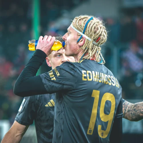 Na zdjęciu widoczny jest piłkarz z warkoczami, który pije napój izotoniczny podczas meczu, a obok niego stoi jego kolega z drużyny.