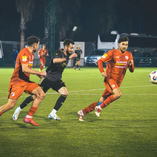 Na zdjęciu widać zawodników w różnych strojach grających w piłkę nożną na boisku, z jednym z graczy w czarnym stroju próbującym przejść obok dwóch przeciwników w czerwonych koszulkach.