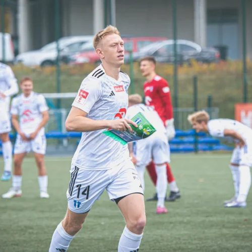 Na zdjęciu widać piłkarza w białym stroju, który trzyma w rękach kartkę, podczas gdy jego koledzy z drużyny przygotowują się do meczu na boisku.