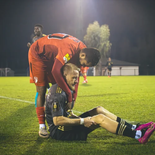 Dwóch piłkarzy na boisku, gdzie jeden z nich pochyla się nad siedzącym na trawie kolegą, wykazując troskę i wsparcie.