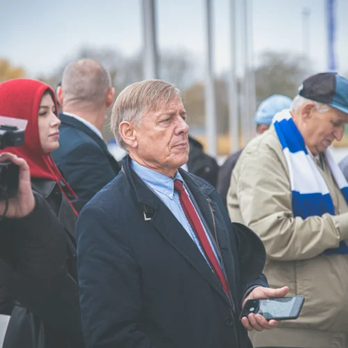 Grupa osób stoi na zewnątrz, jedna z nich trzyma telefon, a w tle widać flagi i zamglone niebo.