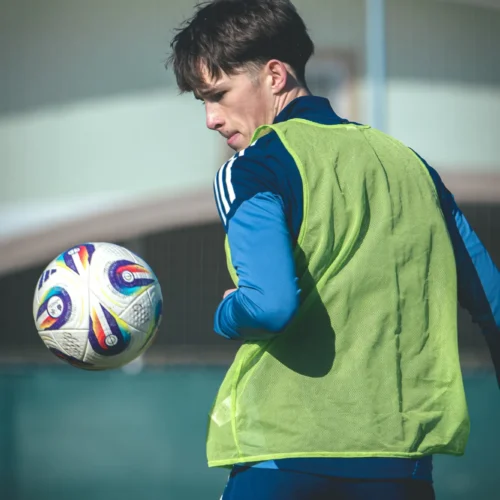 Młody piłkarz w zielonej kamizelce trenuje na boisku, oglądając w powietrzu piłkę nożną.