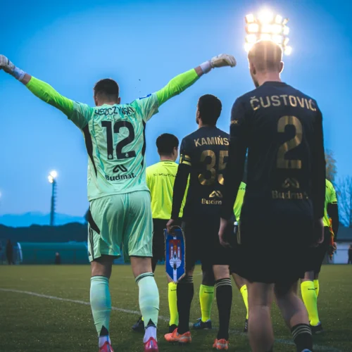 Na zdjęciu widać grupę piłkarzy wychodzących na boisko, w tym bramkarza w jasnej koszulce z numerem 12, w atmosferze zachodzącego słońca.