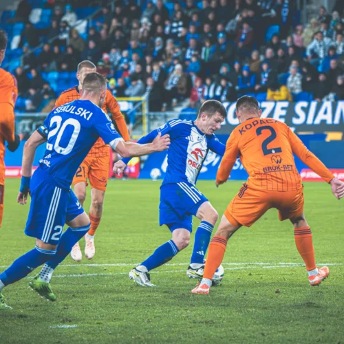 Na zdjęciu widoczna jest intensywna sytuacja na boisku podczas meczu piłkarskiego, w której zawodnik w niebieskim stroju próbuje przejść obok dwóch rywali w pomarańczowych strojach.