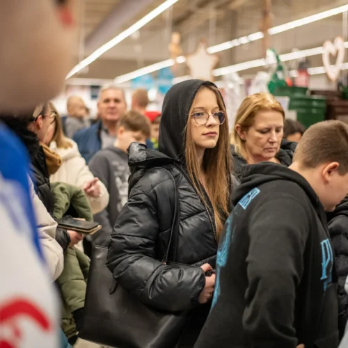 Na zdjęciu widać grupę ludzi stojących w sklepie, z wyraźnym ujęciem młodej kobiety w czarnej kurtce i okularach, która wydaje się zamyślona w tłumie.