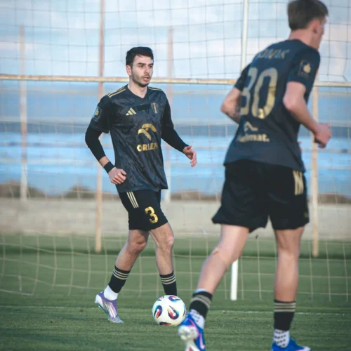 Dwaj piłkarze w czarnych strojach trenują na boisku, z piłką w centrum uwagi.